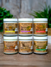 Five jars of Whipped Shea Butter from Moon Artisan Soap on a wooden surface with a blurred green plant background.