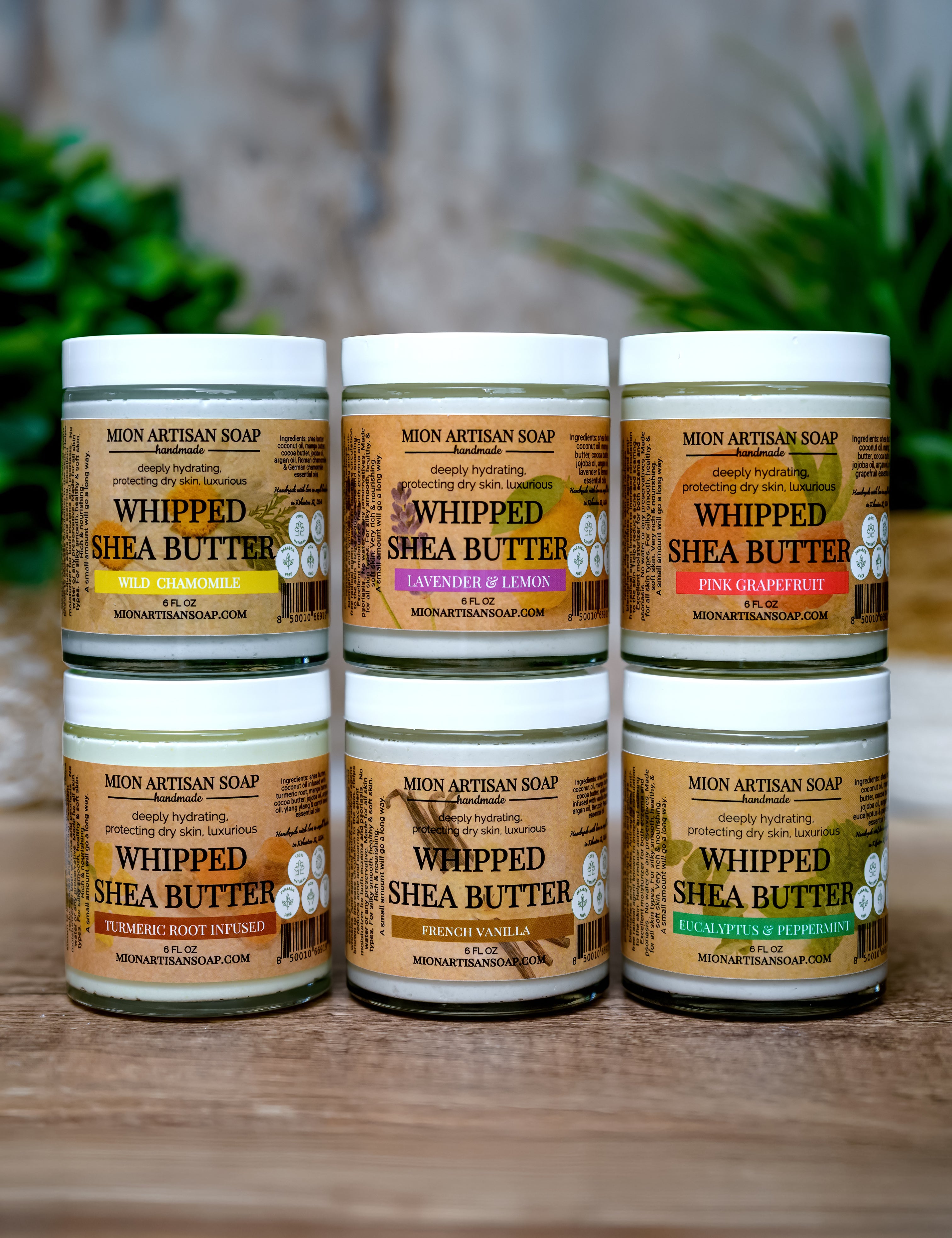 Five jars of Whipped Shea Butter from Moon Artisan Soap on a wooden surface with a blurred green plant background.
