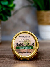 Hand balm container labeled 'Pine & Cedar' on a wooden surface with a blurred natural background.