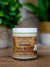 Jar of Mion Artisan Soap Whipped Shea Butter on a wooden surface with a blurred green plant background.
