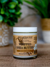 Jar of Mion Artisan Soap Whipped Shea Butter on a wooden surface with a blurred green plant in the background