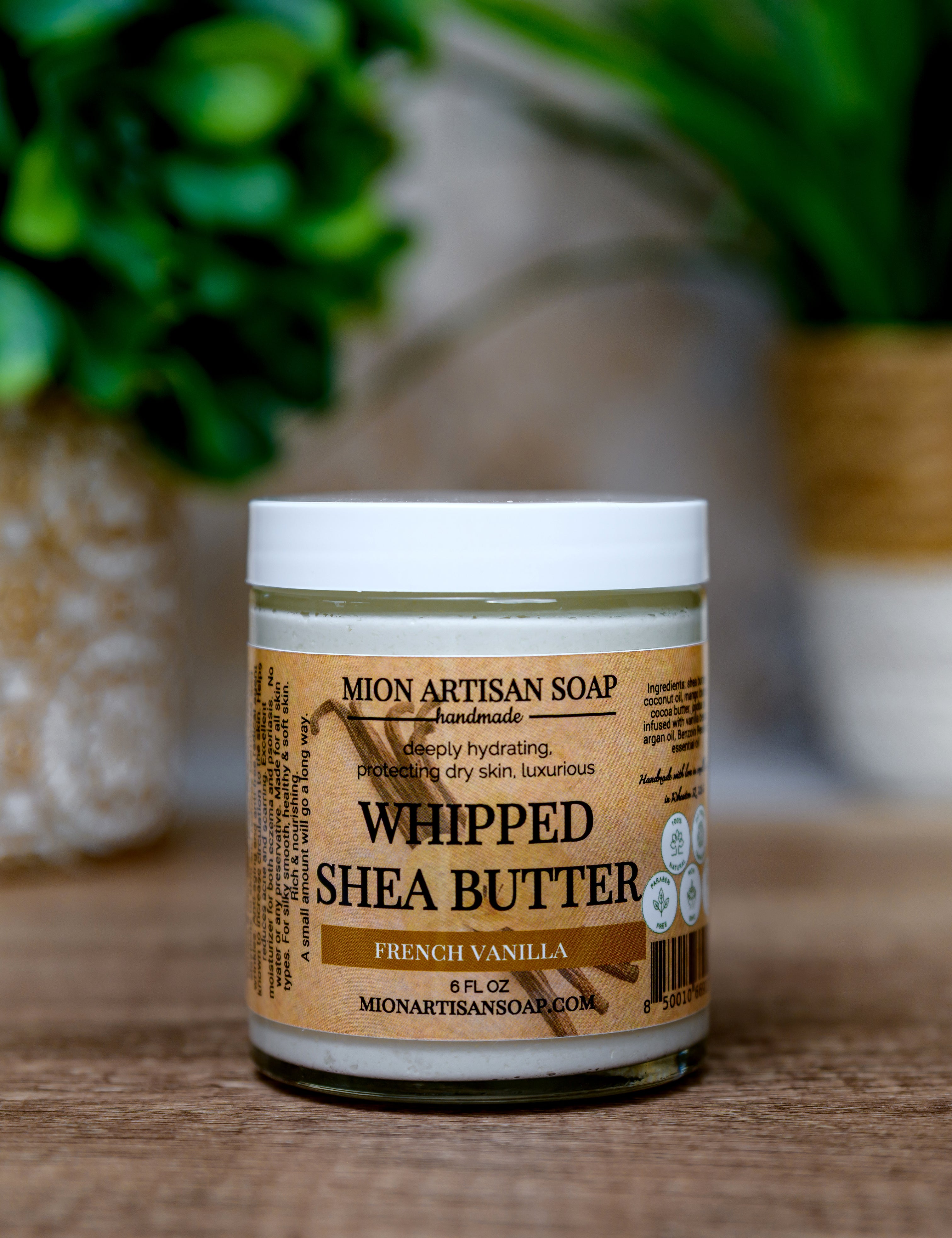 Jar of Mion Artisan Soap Whipped Shea Butter on a wooden surface with a blurred green plant in the background